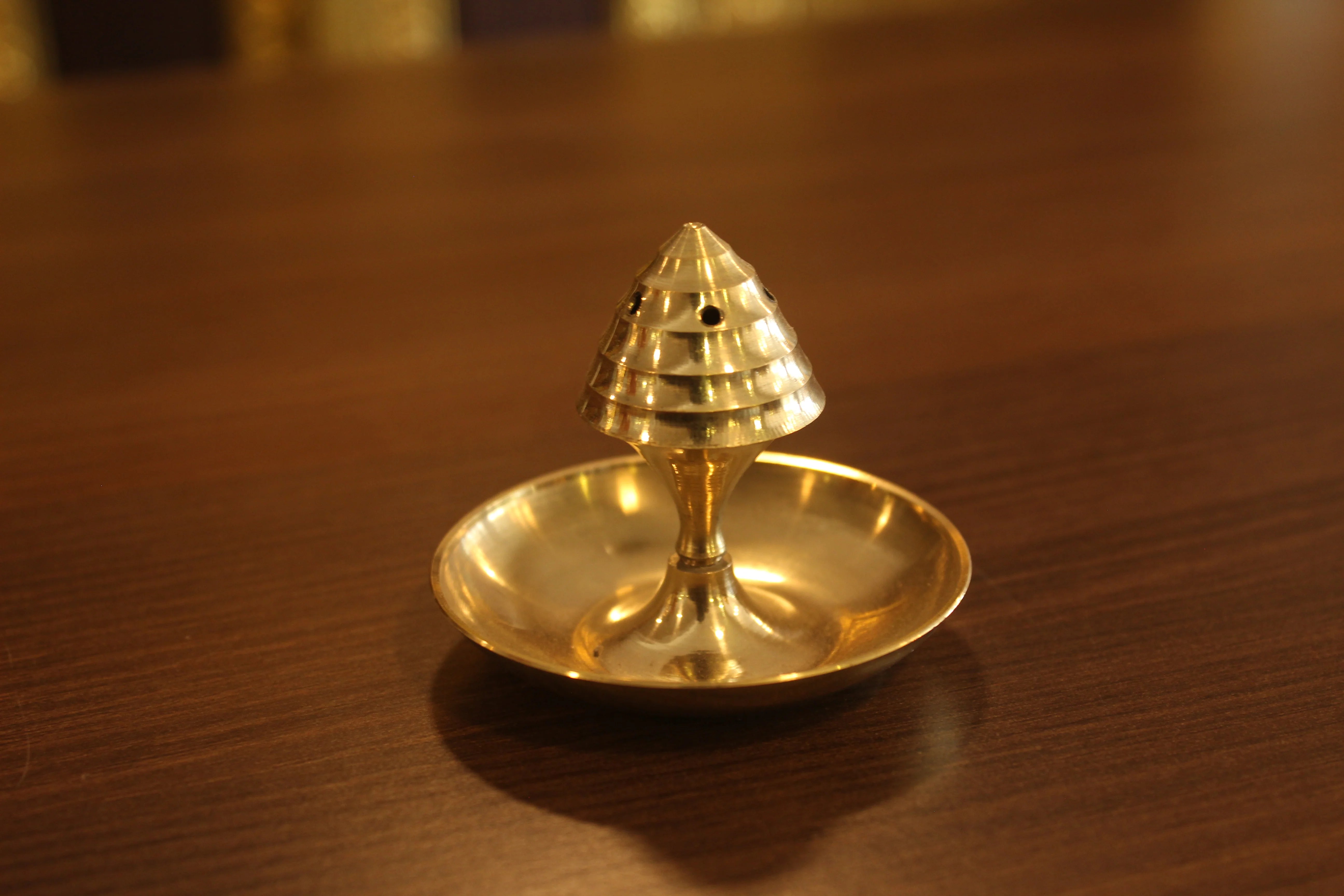 A brass agarbatti stand with a conical shape and a floral pattern on the top, placed on a wooden surface.